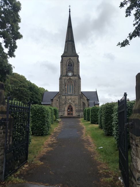 The image depicts a pathway leading to a historic stone church with a tall, pointed spire topped with a cross. The church features a large central clock on the facade and arched windows. Flanking the