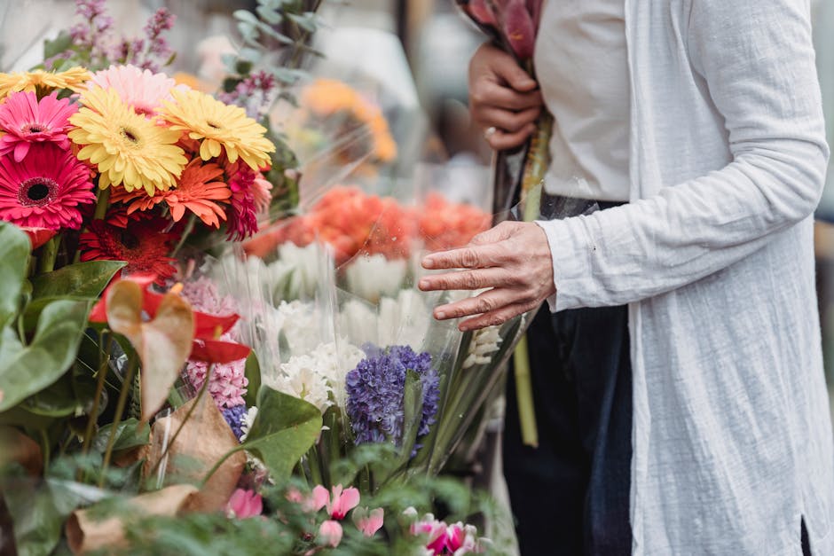 A person wearing a light gray cardigan and dark trousers inspects a vibrant display of fresh flowers at a florist, with bouquets featuring pink, yellow, purple, and white blooms, wrapped in clear plas