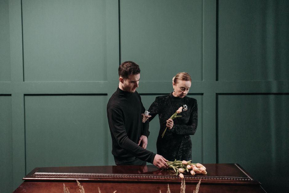 A young man and an older woman are standing at a dark wooden table, with the young man reaching out to touch a bouquet of pale pink roses arranged in a line on the table. The woman, dressed in a black