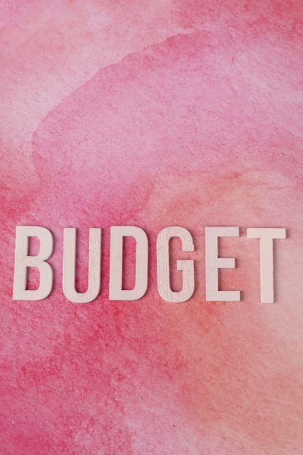 A close-up image featuring the word 'BUDGET' spelled out in bold, white capital letters on a textured pink watercolor background. The background showcases various shades of pink, from light to darker