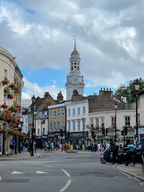 A bustling street scene in daytime with a historic white clock tower featuring a gold weather vane at the top, surrounded by traditional UK high-street buildings. The shops display colorful window dis