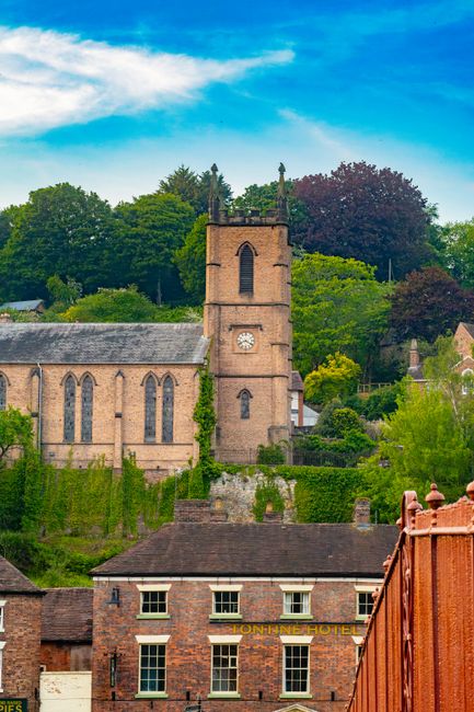 This image features a picturesque scene of a historic brick church with a prominent square clock tower, situated amidst lush green trees and foliage on a bright day with a clear blue sky and some wisp