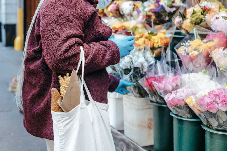 A person wearing a burgundy fleece jacket and blue gloves is selecting flowers at an outdoor flower market, standing next to an array of colorful floral bouquets wrapped in clear paper. The bouquets i